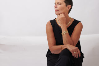 Eine Frau sitzt vor weißem Hintergrund. Am Arm ein goldenes Armband, um den Hals eine goldene Kette.